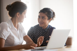 Two female co-workers sitting at desk converse about an issue. A laptop is on the desk.